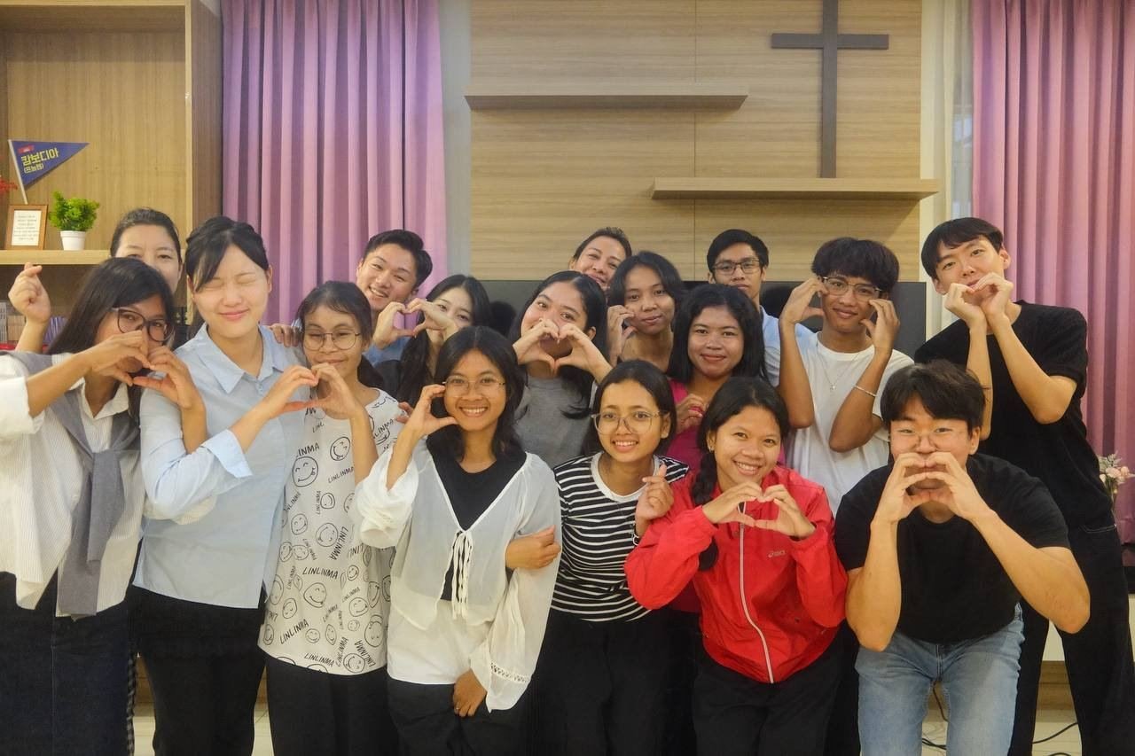 The Phnom Penh group, about twenty young people smiling and making heart hands in a room with a cross on the wall