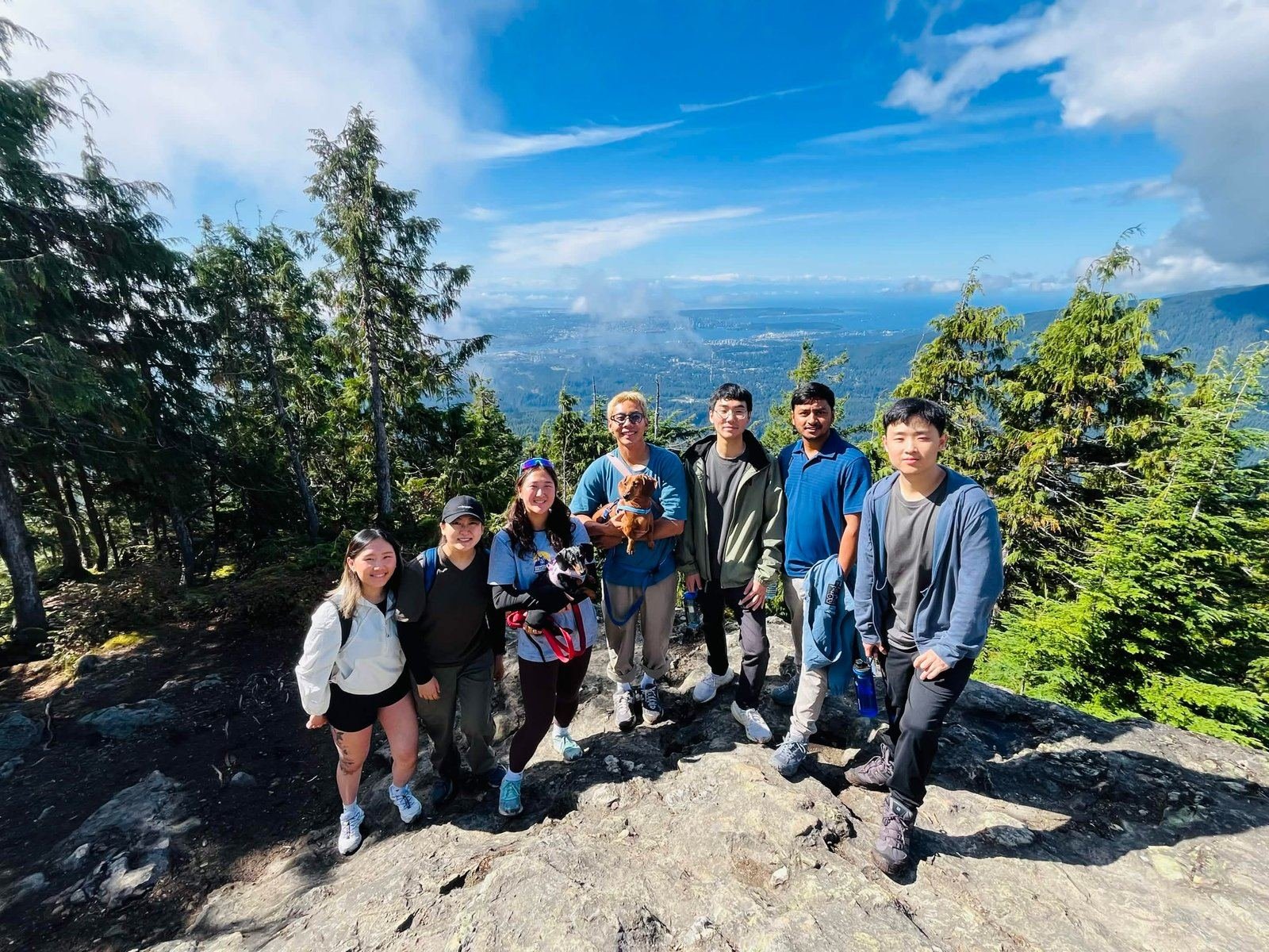 Eight young adults standing together on a rocky mountaintop, evergreen trees around them and the sea and coastline far below under a blue sky