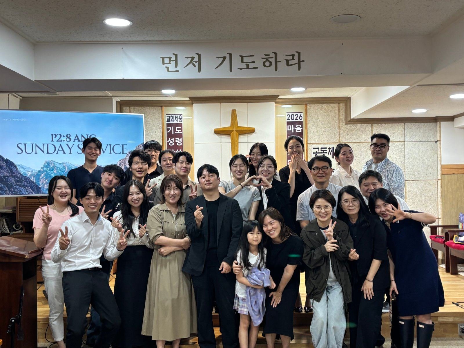 The Seoul congregation gathered at the front of the sanctuary after a Sunday service, a wooden cross behind them and a screen reading P2:8 ANC Sunday Service